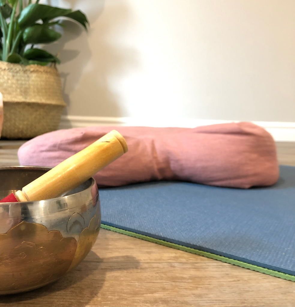 Picture is of a yoga studio. A zoomed view of a meditation cushion, yoga mat, Tibetan signing bowl and house plant. use to advertise weekly yoga classes live, online.